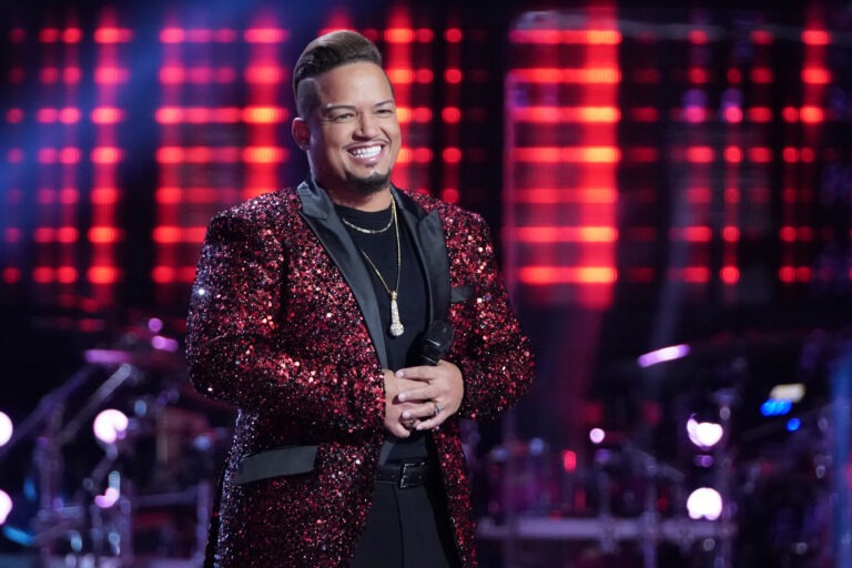 A smiling man in a glittering red jacket stands on stage, illuminated by colorful lights, holding a microphone, conveying excitement and energy.